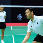 배드민턴 개인 레슨 vs 그룹 레슨 - A focused scene on a brightly lit, modern indoor badminton court. A professional male badminton coac...
